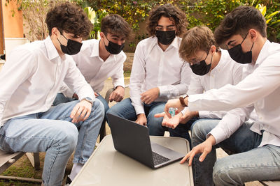Young men wearing mask using hand sanitizer while sitting by laptop outdoors