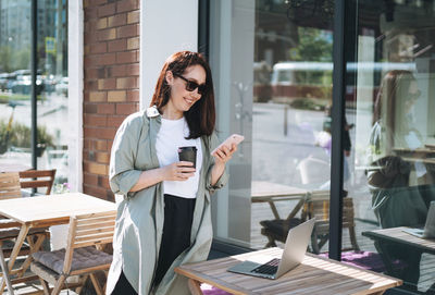 Adult smiling brunette business woman in stylish shirt working on laptop using mobile phone in cafe 