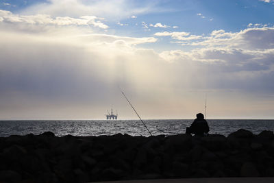 Silhouette of fisherman on rocks with ocean oil rig in the background