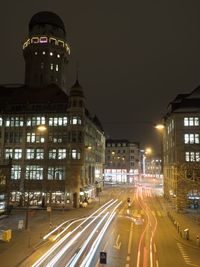 Light trails on city street by buildings against sky at night
