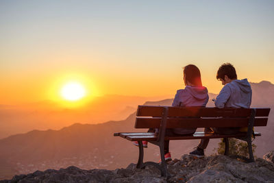 Rear view of couple sitting on bench against sky during sunset