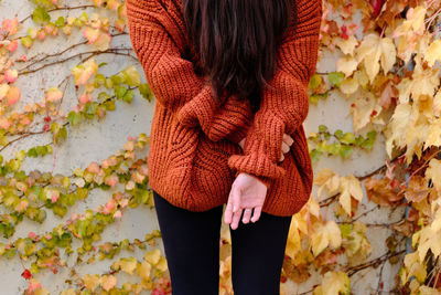 Cropped back view of unrecognizable lonely long haired brunette in red knitted sweater standing against concrete wall covered by colorful ivy plants in autumn day