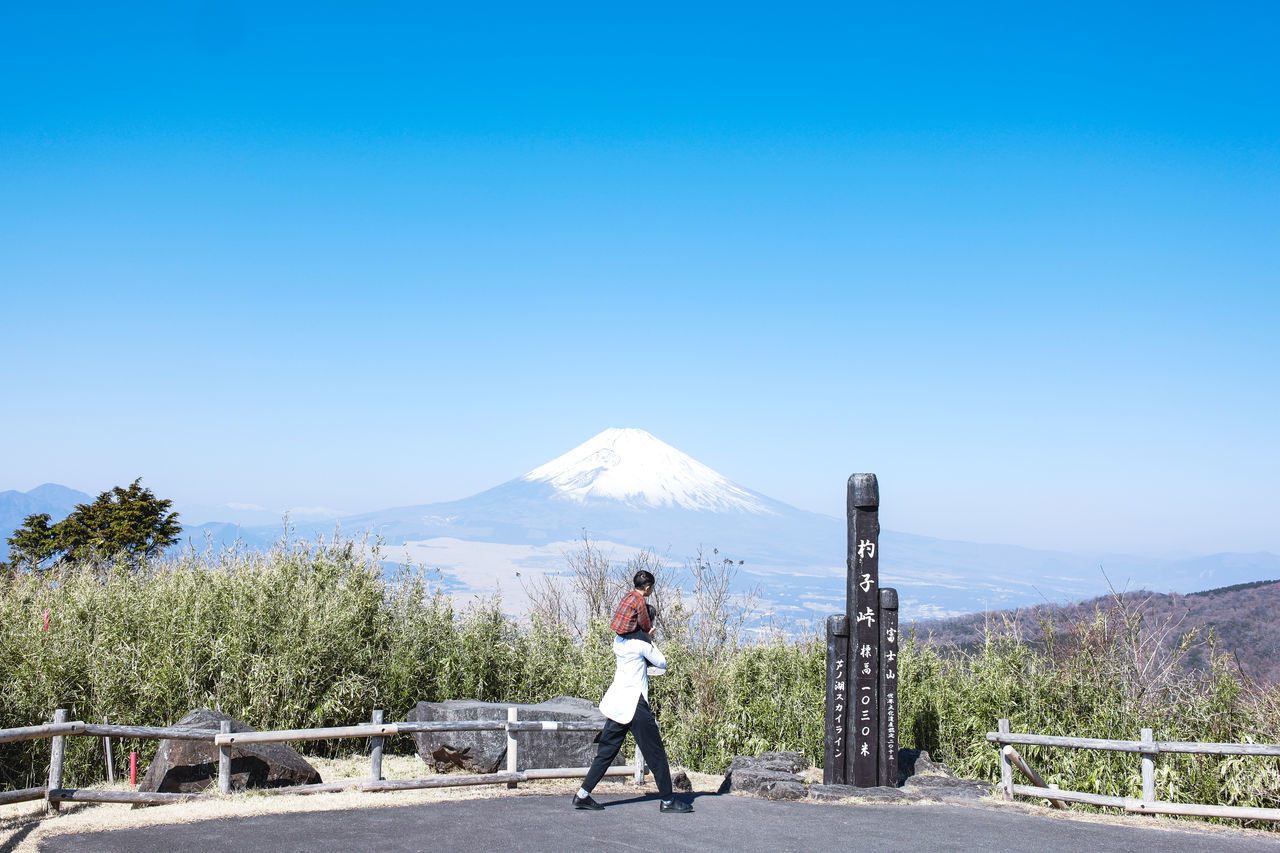 mountain, sky, one person, full length, nature, clear sky, blue, day, scenics - nature, sunny, beauty in nature, leisure activity, adult, copy space, landscape, road, transportation, men, travel, environment, sunlight, plant, rear view, tree, mountain range, outdoors, standing, lifestyles, travel destinations, sports, vacation, tranquil scene, tranquility, non-urban scene, casual clothing, young adult