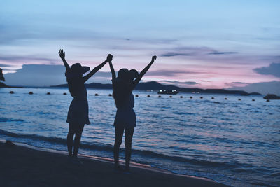 Silhouette female friends with arms raised standing at beach against sky