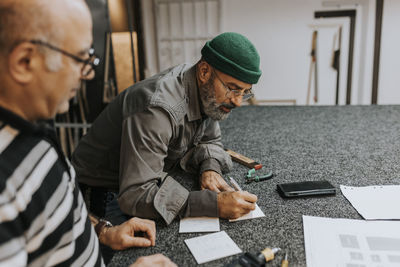 Craftsman wearing knit hat writing by male coworker at workbench