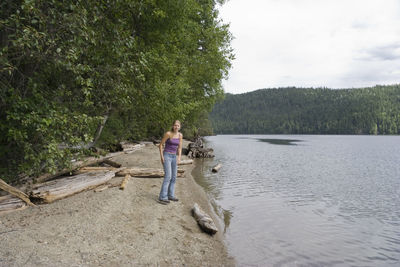 Young woman on lakeshore by trees against sky