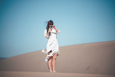 Full length of woman photographing mobile phone while standing at desert against clear sky during sunny day