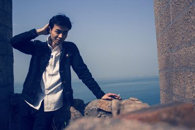 Portrait of young man standing by rock against sea and clear sky