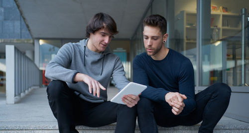 Front view of two young men talking sitting on a city staircase and lo