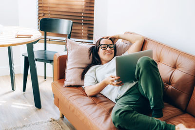 Happy relaxed young beautiful modern brunette lying on sofa using laptop.