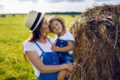 Daughter with mom standing in the field at the sheaf in hats and denim jumpsuits during sunset