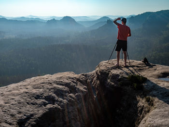 Man shadowing eyes at his big camera on tripod, taking photo of amazing day in hilly landscape