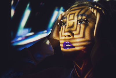 Asian female model with creative makeup standing in studio illuminated by colorful projector lights and looking away while touching head