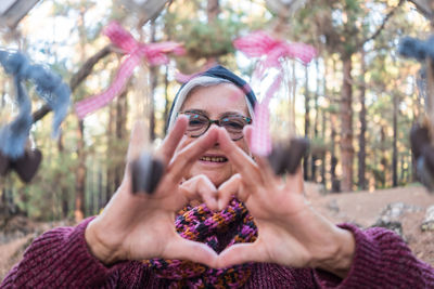 Portrait of senior woman making heart shape by decorations at forest