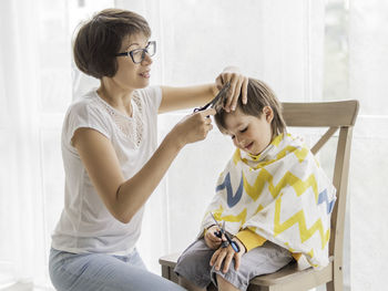 Mother cuts her son's hair by herself. little boy sits, covered with cloth, holds pair of scissors. 