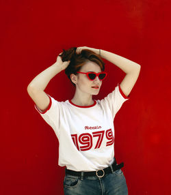 Woman wearing sunglasses and tying hair while standing against red wall