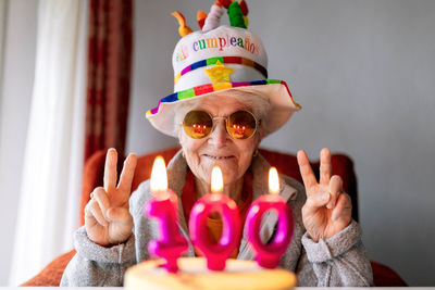 Delighted senior female in funny hat and sunglasses showing v sign gesture with both hands and celebrating birthday with cake with 100 candle