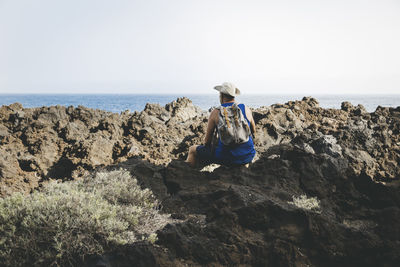 Rear view of woman sitting on rock at beach against clear sky