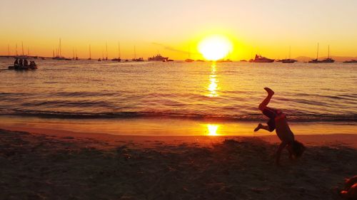 Shirtless boy performing handstand at beach against sky during sunset