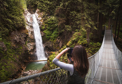 Woman photographing waterfall while standing on footbridge at forest