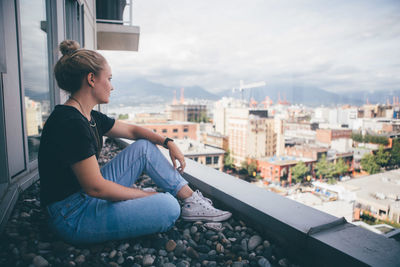 Young woman sitting on retaining wall against city