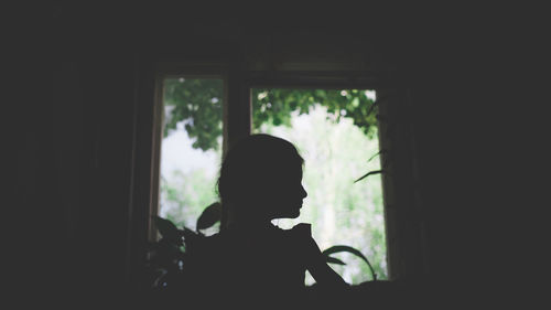 Rear view of silhouette woman looking through window at home