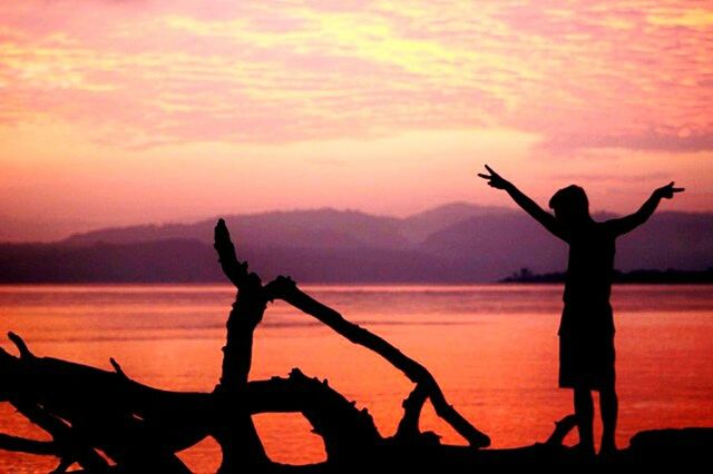 sunset, silhouette, sky, leisure activity, lifestyles, orange color, water, arms raised, sea, scenics, standing, cloud - sky, beauty in nature, tranquil scene, arms outstretched, enjoyment, full length, tranquility