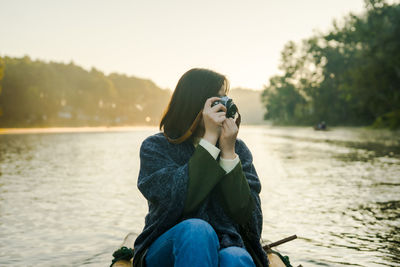 Full length of woman photographing by lake against sky