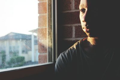 Thoughtful young man sitting by window against brick wall at home