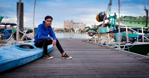 Portrait of man sitting on pier against sky by moored boats 