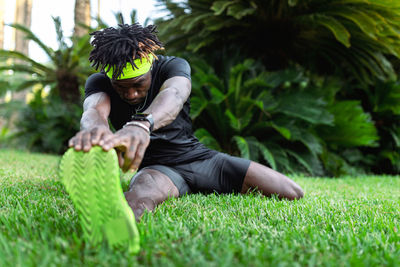 Young african american sportsman in stylish activewear sitting on green grass and doing stretching exercises after running in park