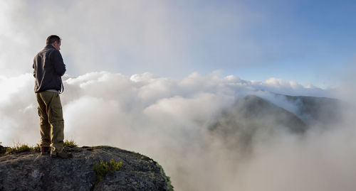 Man standing on cliff by fog covered mountains against sky