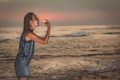 Optical illusion of woman eating sun at beach against sky during sunset