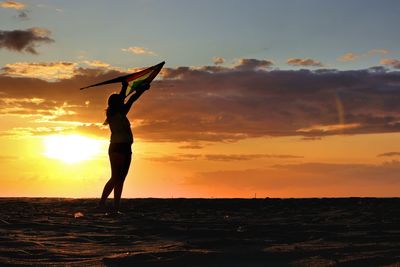 Silhouette girl holding kite while standing at beach against sky during sunset