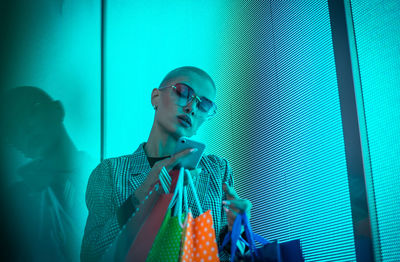 Low angle view of lesbian woman using smart phone holding shopping bags standing against abstract backgrounds