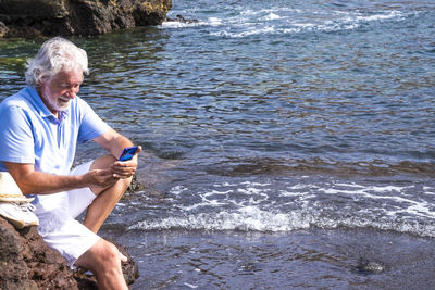 Smiling senior man using smart phone sitting on rock by sea
