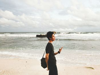 Side view of young man gesturing while standing on beach against sky