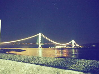 Illuminated suspension bridge over river against sky at night