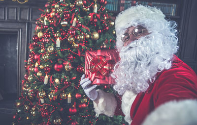 Man wearing santa claus costume with gift by christmas tree at home