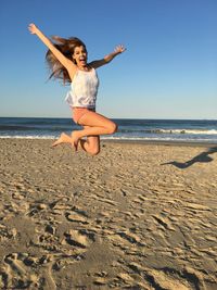 Full length of cheerful young woman with arms raised jumping on shore at beach