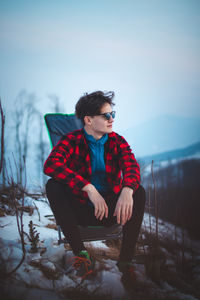 Handsome teenager aged 15-20 in a plaid red-black shirt and sunglasses on a chair by the tent. 