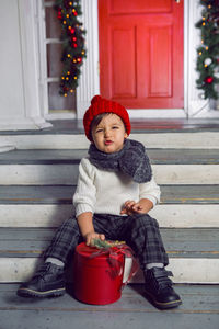 Child boy in a white sweater and a red knitted hat sits on the steps with snow at home at christmas