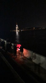 People on illuminated building by sea against sky at night