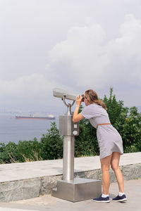 Young woman using stationary binoculars on a warm summer day on a hill against the background