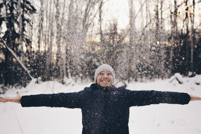 Snow falling on man with arms outstretched standing against trees during sunset