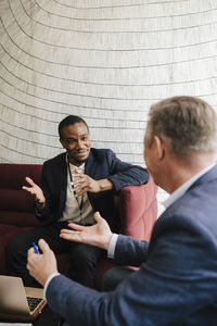 Male colleagues gesturing while discussing together sitting on sofa during networking event