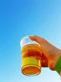 Cropped hand holding beer against clear blue sky