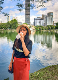 Young woman drinking water while standing against river