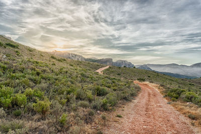 Dirt road crossing a mountainous landscape at sunset with a cloudy sky