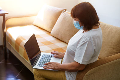 Serious woman in protective mask uses laptop, checks email on internet while